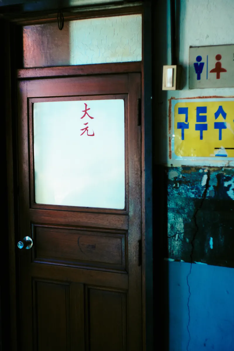 Wooden door with a frosted glass window and a red character, beside a yellow sign in a market corridor