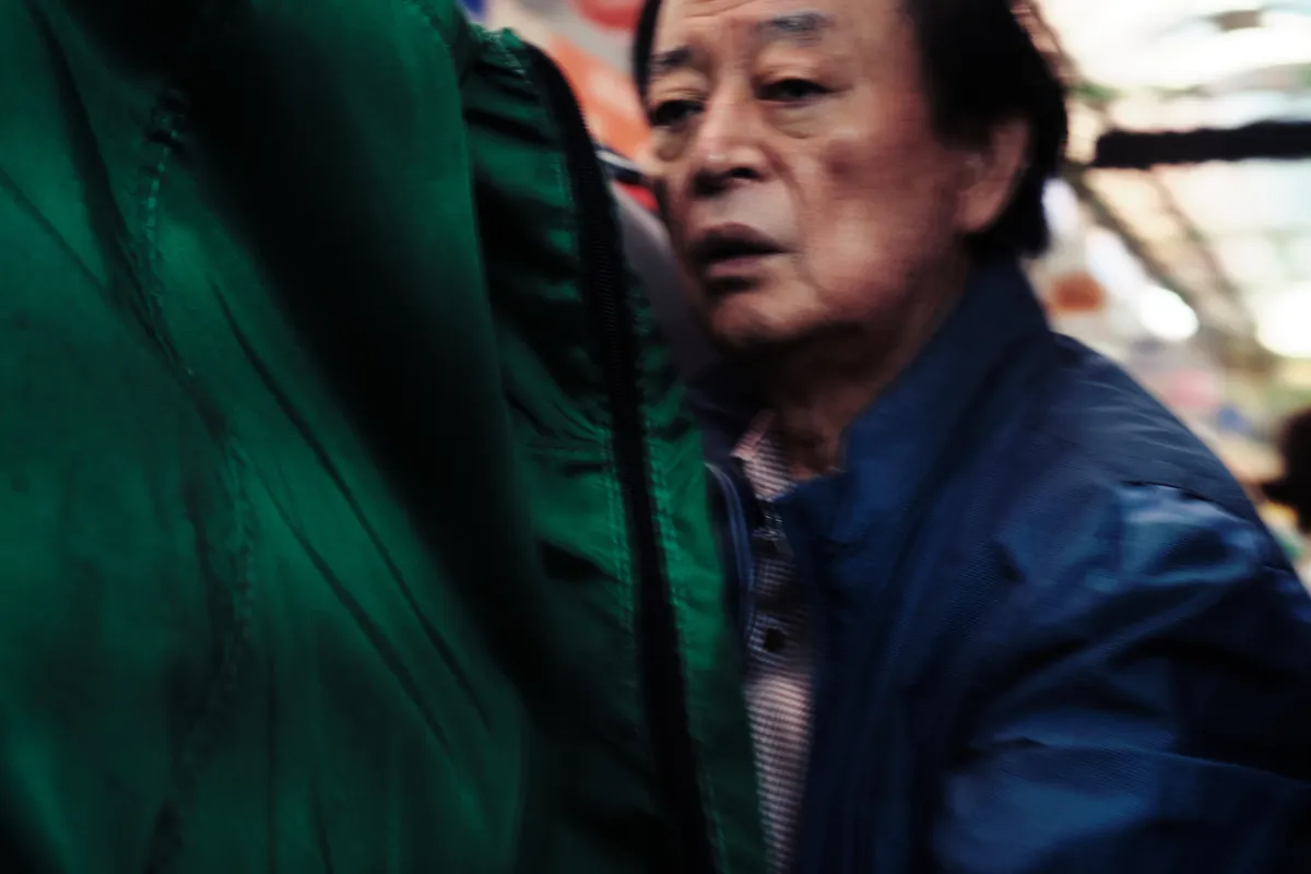 Older man in a blue jacket beside hanging green clothes in a market