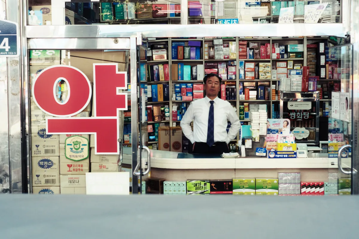 Pharmacist in a white shirt and tie standing behind the counter of a small shop under a red 약 sign