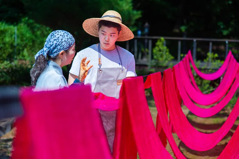 Haram Jackson Park in a straw hat talking with a woman among lines of red safflower-dyed silk hanging outdoors